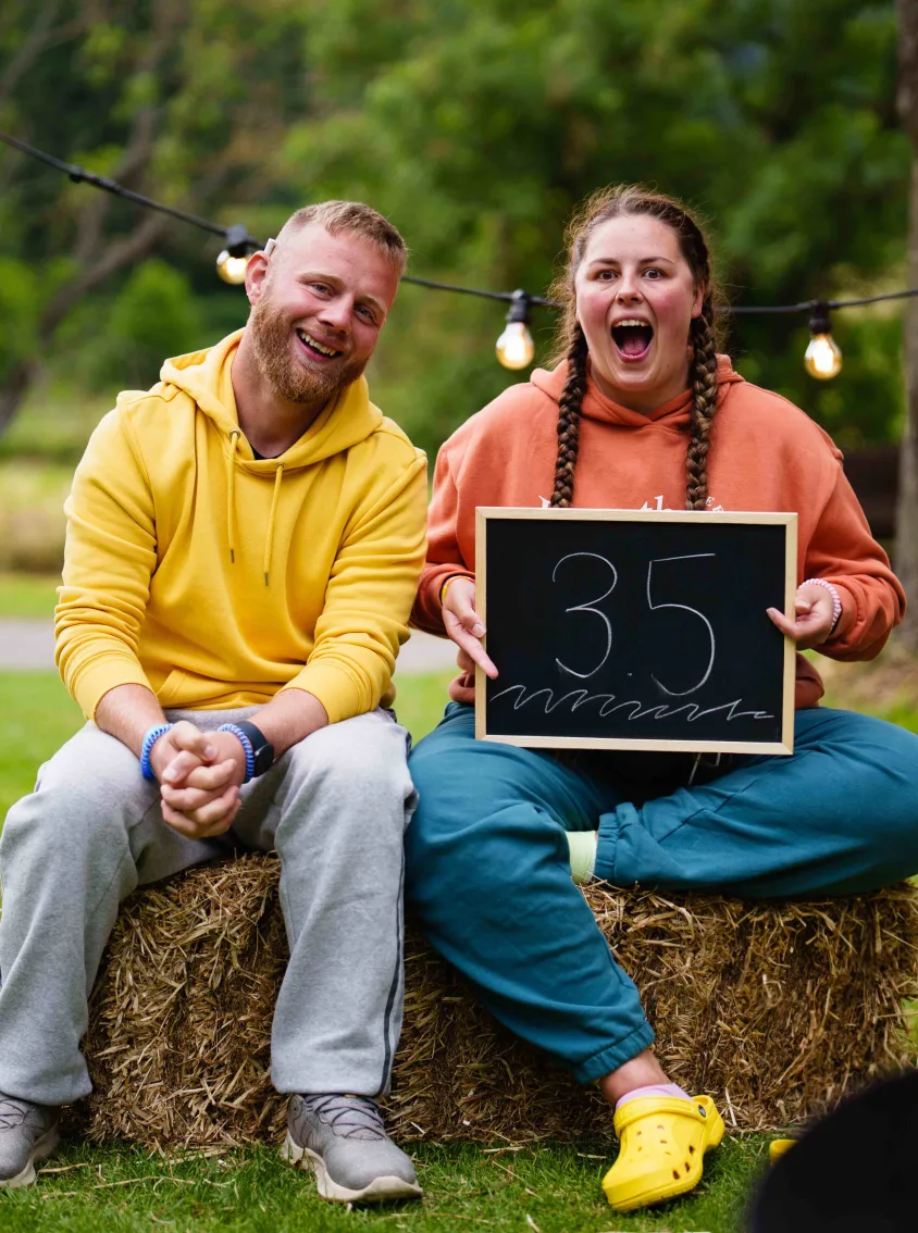 man and woman sat on a hay bail in a field holding a sign with 35 written on it